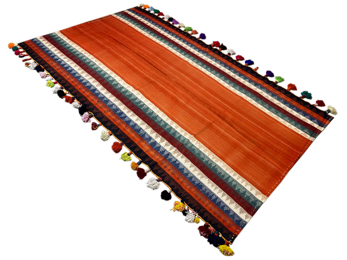 Tapis Jajim déployé en diagonale : terracotta, bordure rayée, pompons de différentes couleurs