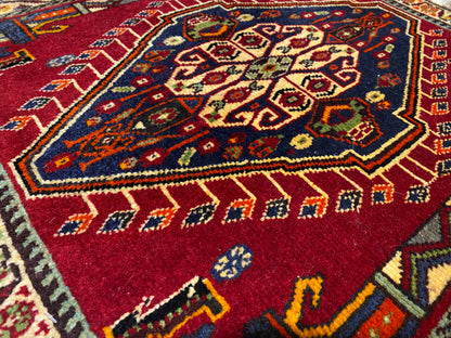 Gros plan sur un tapis Poshti (59×61 cm) : motif intérieur rouge orné de motifs colorés et d’une bordure