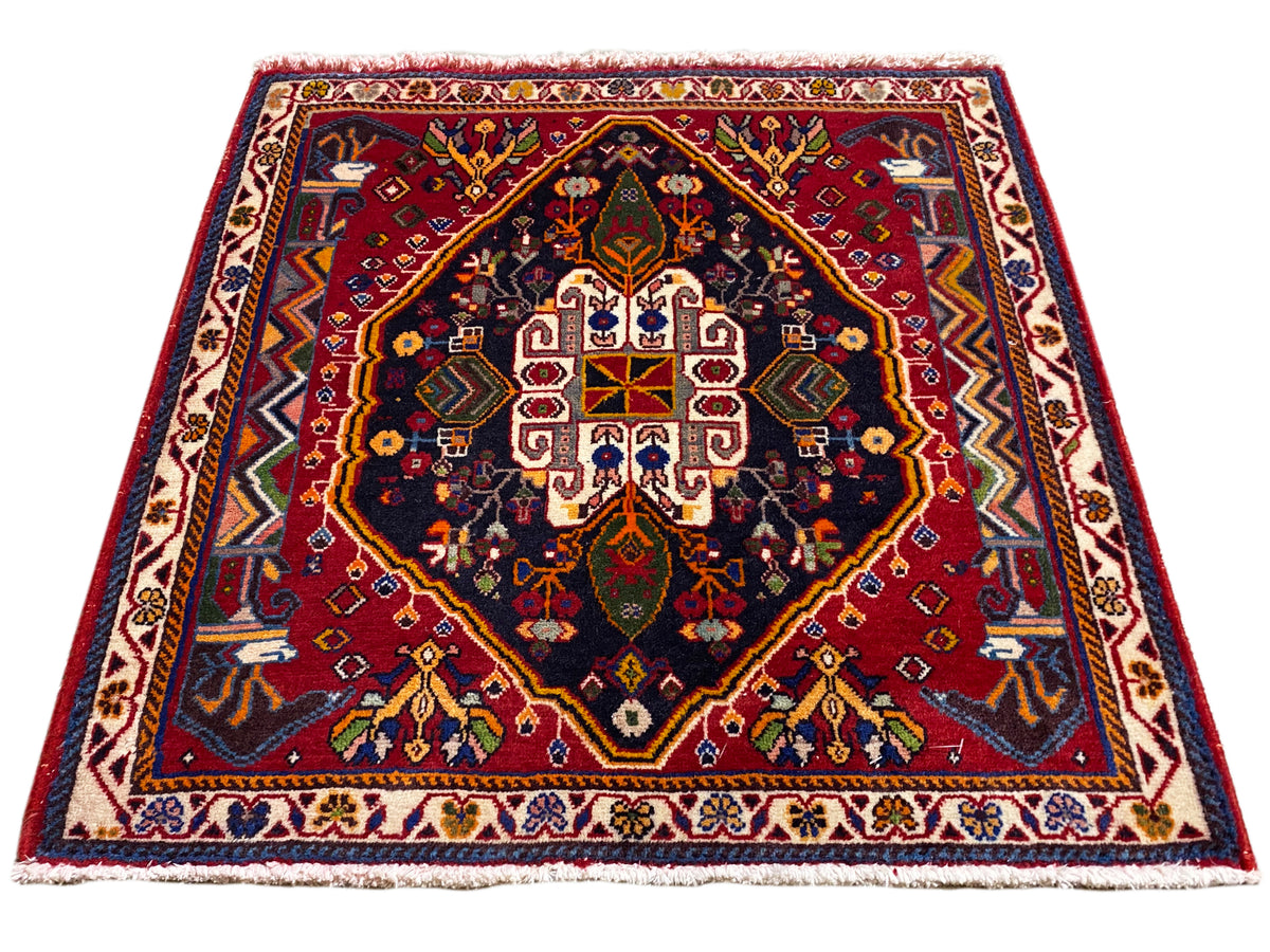Tapis Poshti (67×67 cm) en vue oblique : bordure, centre rouge, bleu foncé